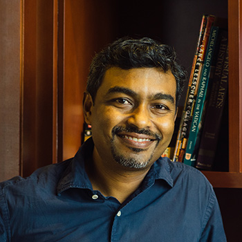 A headshot of smiling Ajay Sriram Mathuru who has short black hair, wearing a blue shirt. There are bookshelves in the background.