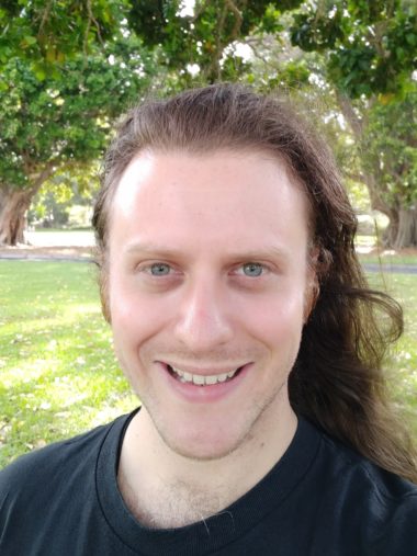 A headshot of smiling David Andrew Smith who has long brown hair and is wearing a black shirt. There is greenery in the background.