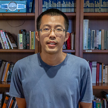 A headshot of smiling Liuchun Deng who has short black hair, wearing glasses with a transparent frame and a blue long-sleeve shirt. He is standing in front of a large bookshelf.