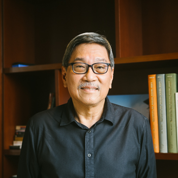 A headshot of smiling Ho Kong Chong who has short black-grey hair, wearing a black shirt and black-rimmed glasses. He is standing in front of a large bookshelf.