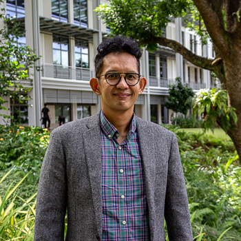 A headshot of smiling Nur Amali Ibrahim who has short, curly black hair, wearing glasses with black frame and a grey wool suit with green, blue, and red flannel shirt. He is standing in the Yale-NUS Campus Green with the Yale-NUS library building in the background.