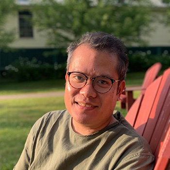 A headshot of smiling Lawrence Lacambra Ypil who has short grey hair. He is wearing glasses with a brown frame, and a green t-shirt. He is sitting on a red deck chair in a courtyard with a house and a tree in the background.