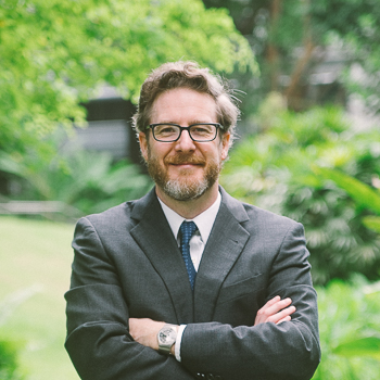 A headshot of smiling David M. Post who has short brown hair, wearing a black suit and black-rimmed glasses. He is posing in front of lush greenery and has his arms crossed in front of him.