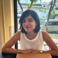 A headshot of smiling Sneha Annavarapu who has short black hair of shoulder length in bob cut, wearing a white sleeveless top, a square watch on her arm, a necklace, and a flower-shaped ring. She is sitting at a restaurant with her hands placed on the table. In the background, there is a window that overlooks a house, trees, and a car.