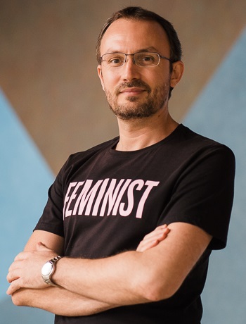 A headshot of smiling Stanislav Presolski who has short brown hair, a mustache, and a beard, wearing a black “Feminist” t-shirt. He is posing by crossing his hands over his chest, standing at the Yale-NUS Performance Hall foyer, in front of a wall drawing.