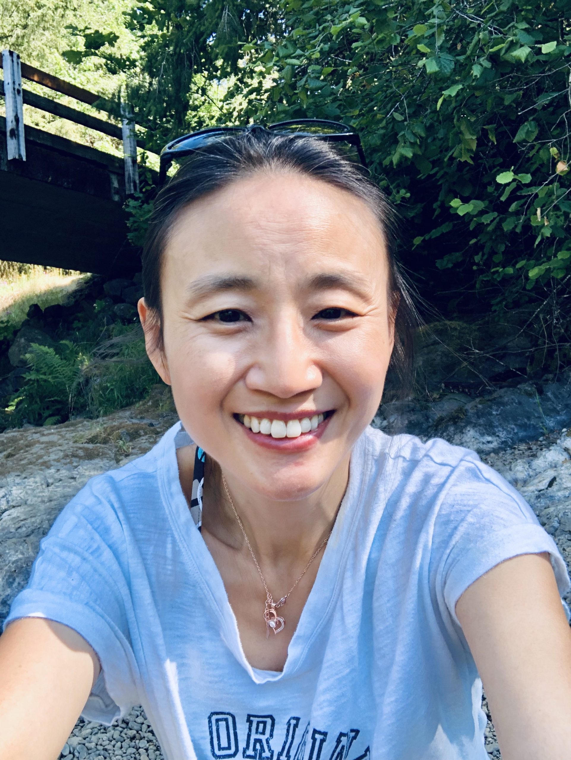 A headshot of smiling Yin Chang who has black hair in a ponytail. She is wearing sunglasses on top of her head, a gold necklace with a pearl, and a grey T-shirt. She is sitting on a rock with green plants and a bridge in the background.