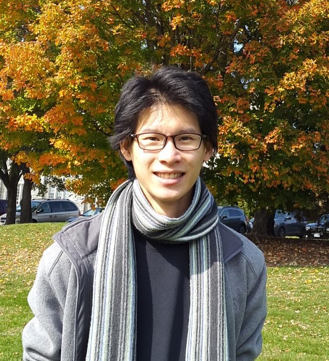 A headshot of smiling Yunus Prasetya who has black hair. He is wearing glasses with a black frame, a grey jacket with a black shirt, and a grey and white striped scarf. In the background there are trees with green and yellow leaves.