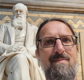 A headshot of smiling Todd Jared LeVasseur who has brown hair, a mustache, and a grey beard. He is wearing black-framed glasses and is posing in front of a white marble statue of Charles Darwin.