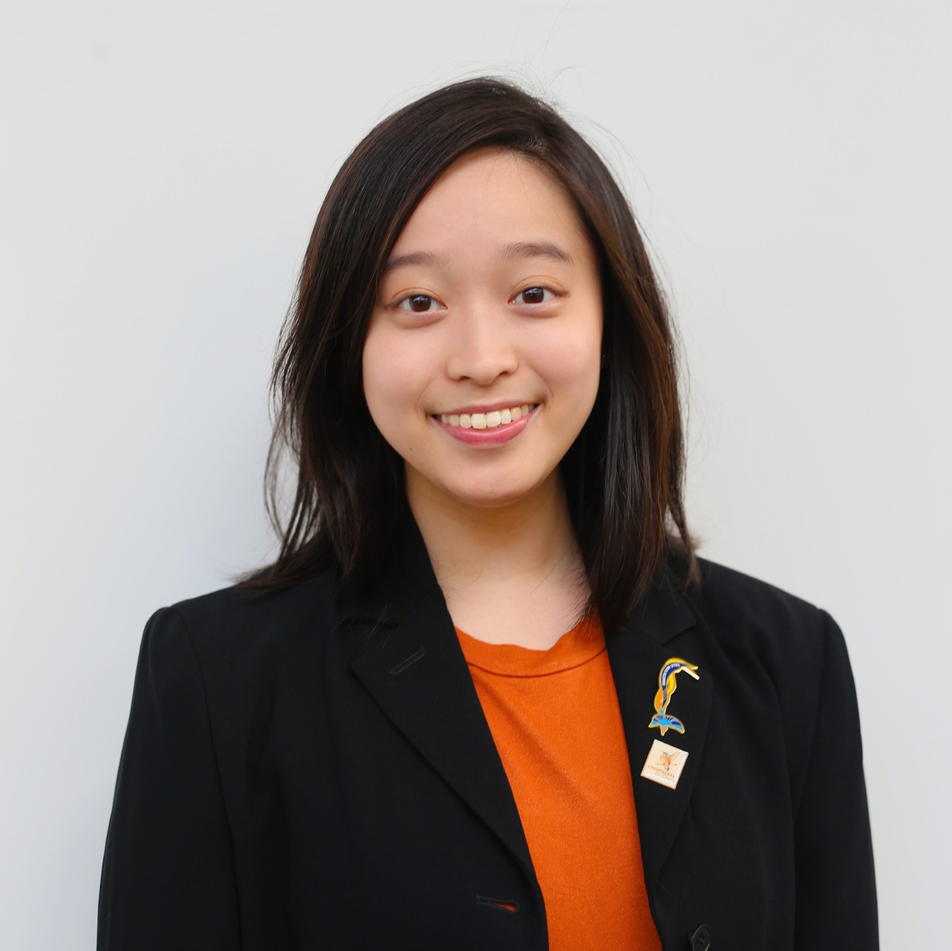 A headshot of smiling Sherice Ngaserin Ng Jing Ya who has black hair of shoulder length. Sherice is wearing an orange shirt and a black blazer, on the left lapel are Yale-NUS pins for alumni and Kingfishers for Consent.